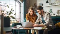 Un jeune couple, assis sur leur canapé dans leur appartement, utilise un ordinateur portable pour remplir formulaire, des feuilles de papiers sur la table.