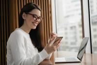 Une femme souriante consulte son smartphone devant son ordinateur portable