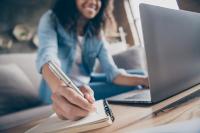 Photo recadrée avec mise au point flou d’une jeune femme qui écrit au stylo dans un carnet de note assise dans un canapé