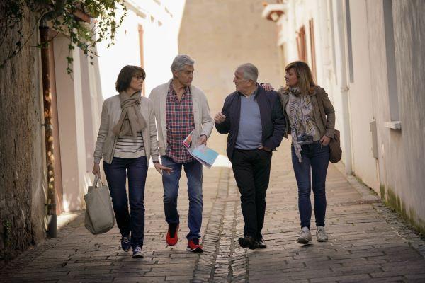 Deux couples de senior discutant et marchant de face dans une rue piétonne pavée ensoleillée.