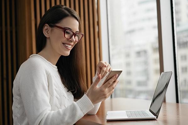 Une femme souriante consulte son smartphone devant son ordinateur portable